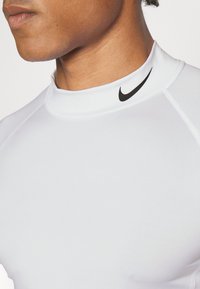 Bjela atletska majica s visokim ovratnikom, s crnim Nike logotipom. Glatka tekstura i prilagođen dizajn poboljšavaju performanse.