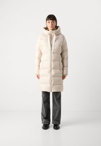 Zachte beige lange jas met een rits aan de voorkant, verstelbare koordkap en grote zijzakken. Draagt over donkergrijze flared jeans en zwarte schoenen.