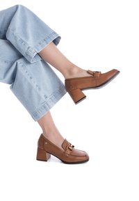 Unos zapatos de tacón de cuero marrón presentan una puntera redondeada, una hebilla decorativa y un tacón cuadrado con una suela negra contrastante, combinados con unos pantalones vaqueros de corte recto y de tono azul claro.