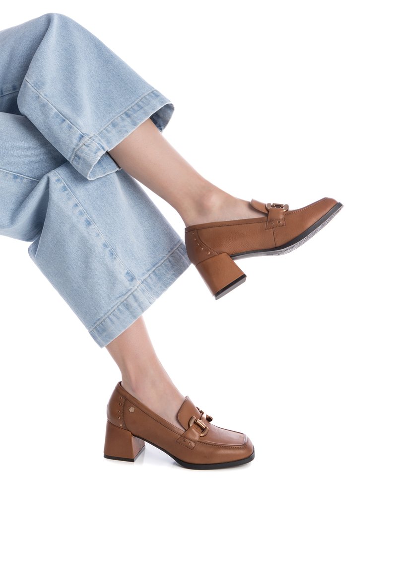 Unos zapatos de tacón de cuero marrón presentan una puntera redondeada, una hebilla decorativa y un tacón cuadrado con una suela negra contrastante, combinados con unos pantalones vaqueros de corte recto y de tono azul claro.