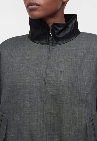 Personne portant une veste grise texturée avec un col montant en cuir noir zippé autour du cou, montrant les lèvres et le menton.