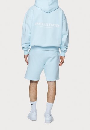 Person trägt hellblauen Hoodie und passende Shorts mit weißem Schriftzug „PEGADOR“ auf dem Rücken und weiße Sneakers, steht mit dem Rücken zur Kamera.