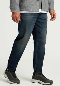 Slim fit sötét farmernadrág enyhe kopással, fekete sportcipővel, fehér talppal és szürke kiegészítőkkel párosítva.
