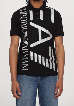 Mann trägt ein schwarzes Emporio Armani Poloshirt mit großem weißem grafischem Text und Formen, kombiniert mit blauen Jeans.