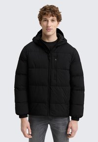 Ung mand med krøllet hår iført en sort hættejakke og grå jeans, der står op ad en lys grå baggrund.