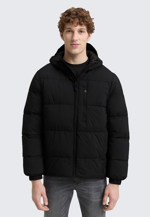 Young man with curly hair wearing a black hooded puffer jacket and gray jeans standing against a light gray background.