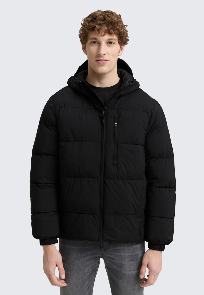 Ung mand med krøllet hår iført en sort hættejakke og grå jeans, der står op ad en lys grå baggrund.