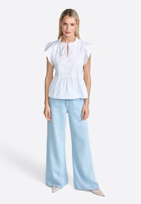 White peplum top with ruffled sleeves, keyhole neck, paired with wide-leg light blue jeans and pointed beige heels on a neutral background.