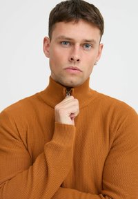 Ráncos, barna, cipzáras pulóver magas gallérral. Texturált anyag és szűkített mandzsetták jellemzik. A modellen a cipzár látható.