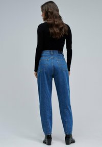 Vaqueros azul de tiro alto fabricados en denim. Presentan piernas estrechas, cinco bolsillos y un sutil acabado desgastado. Se llevan con una blusa negra de manga larga.
