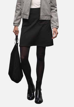 Kobieta w szarej kurtce, czarnej spódnicy, czarnych rajstopach i czarnych botkach, trzymająca w prawej dłoni czarną torebkę.