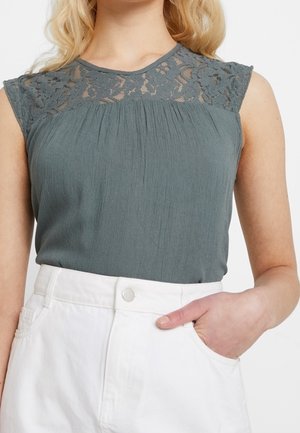 Woman wearing sleeveless gray top with lace yoke and white high-waisted pants, hand resting in pocket, against a plain background.