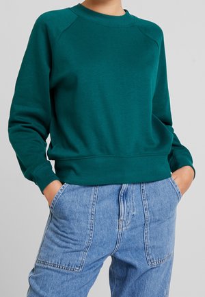 Osoba ubrana w turkusową bluzę z długim rękawem i jasnoniebieskie, wysokowodne dżinsy z rękami w kieszeniach na jednolitym tle.