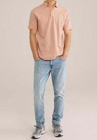 Mężczyzna w różowej koszulce polo z krótkim rękawem, jasnoniebieskich dżinsach i szarych trampkach, stojący z rękami w kieszeniach.