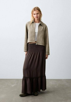 Veste courte en suédine grise imitation, avec fermeture éclair et détails cloutés, associée à une jupe longue marron avec taille smockée et ourlet à volants.