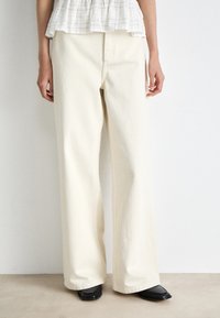 Παντελόνι σε μπεζ απόχρωση με φαρδύ καρέ, με απαλό υλικό και καθαρή γραμμή, διαθέτει εμπρόσθια ραφή και θηλιές για ζώνη, συνδυασμένο με μαύρες σαμπάνες με μυτερή μύτη.