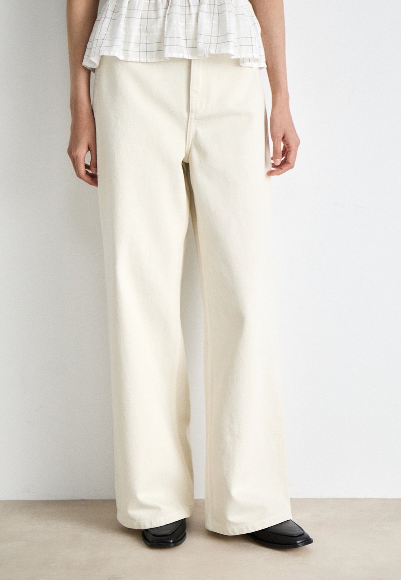 Παντελόνι σε μπεζ απόχρωση με φαρδύ καρέ, με απαλό υλικό και καθαρή γραμμή, διαθέτει εμπρόσθια ραφή και θηλιές για ζώνη, συνδυασμένο με μαύρες σαμπάνες με μυτερή μύτη.