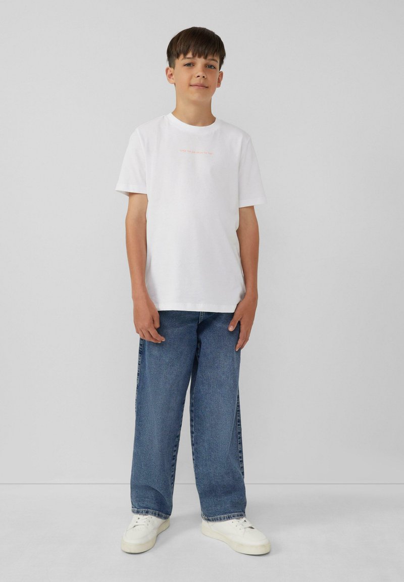 Witte katoenen t-shirt met korte mouwen en kleine oranje tekst. Gecombineerd met losse blauwe denim jeans en witte sneakers. Eenvoudig casual ontwerp.