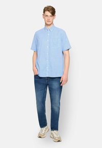 Camisa de botones de manga corta azul claro con un bolsillo en el pecho, combinada con jeans azul oscuro y zapatillas beige, sobre un modelo que se encuentra frente a un fondo blanco.