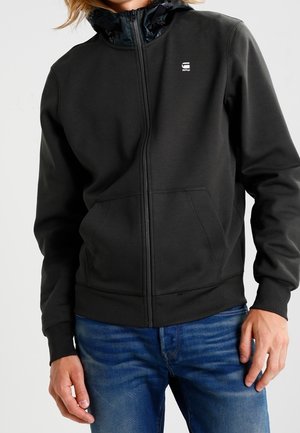 Czarna bluza z kapturem zapinana na zamek, z teksturowanym kapturem, kontrastową podszewką oraz przednimi kieszeniami. Wykonana z gładkiego, miękkiego materiału. Na piersi znajduje się logo.