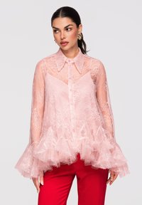 Femme portant un chemisier en dentelle rose transparent avec un ourlet et des poignets à volants, assorti à un pantalon rouge vif, regardant sur le côté les cheveux attachés.