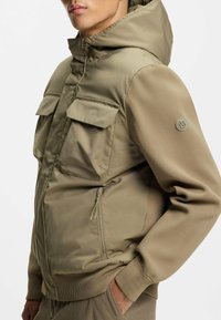 Beige foret jacket med hætte, to frontlommer og ribstrikkede manchetter. Stoffet har en glat overflade med en sidelomme med lynlås.