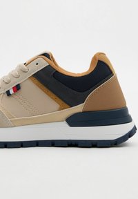 Zapatilla beige con acentos en azul marino y marrón. Presenta un upper texturizado, cuello acolchado y una entresuela blanca con una suela de goma texturizada.