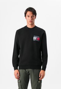Zwarte sweatshirt met een ronde halslijn, ribgebreide manchetten en onderrand. Heeft een logo in blauw, rood en wit op de linkerborst. Gecombineerd met groene cargobroek.