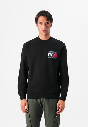 Zwarte sweatshirt met een ronde halslijn, ribgebreide manchetten en onderrand. Heeft een logo in blauw, rood en wit op de linkerborst. Gecombineerd met groene cargobroek.