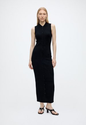 Femme portant une robe longue noire sans manches côtelée boutonnée et des sandales noires à talons ouverts sur fond blanc.