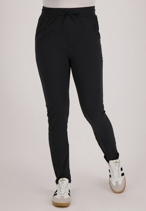 Persoon met zwarte slim-fit broek met een trekkoord in de taille en witte sneakers met zwarte strepen, staand tegen een effen achtergrond.
