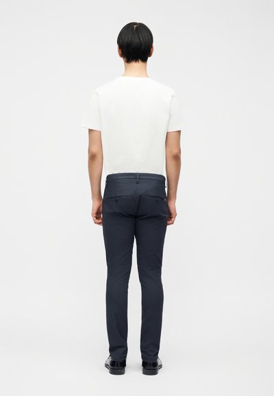 Άνθρωπος που στέκεται με την πλάτη, φοράει λευκό κοντομάνικο πουκάμισο, σκούρο στενό παντελόνι και μαύρα επίσημα παπούτσια σε απλό φόντο.