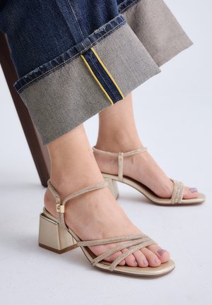 Pies con sandalias beige de tacón bloque con tiras, combinadas con vaqueros oscuros con dobladillo que muestran detalle de costura amarilla.