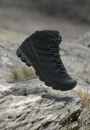 Botte de randonnée noire La Sportiva en l’air au-dessus d’un terrain rocheux avec de petites plantes et un arrière-plan flou.
