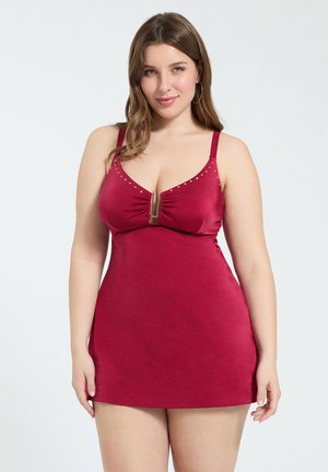 Femme aux cheveux bruns mi-longs, portant une robe rouge ajustée sans manches avec des détails en clous dorés et un fermoir central à l'avant.