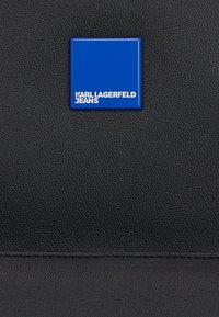 Black textured leather bag featuring a square blue logo badge with "KARL LAGERFELD JEANS" in white text. Simple and modern design.