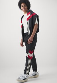 Černý sportovní komplet s červeno-bílým šikmým vzorem, obsahující tři bílé pruhy na každé straně a bílý tričko, doplněný šedými teniskami.