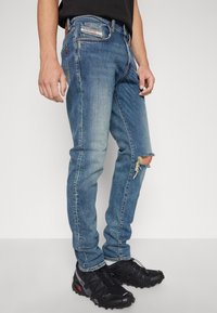 Smal blå jeans i denim, med ett slitigt knä med fransade kanter, framfickor och en subtilt blekt tvätt. Svarta sportskor är på.