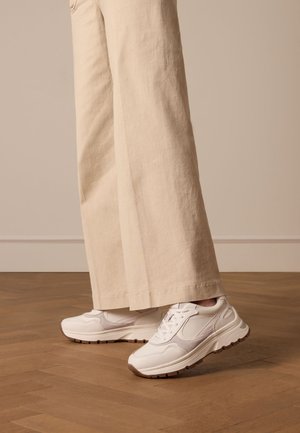 White athletic sneakers with textured overlays, lace-up front, and rubber outsole. Paired with wide, beige linen pants. Wood floor background.
