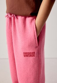 Person wearing pink textured sweatpants with "AMERICAN VINTAGE" text on leg, hand resting inside side pocket, brown top visible.