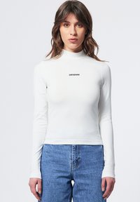 Weißes Langarm-Turtleneck-Oberteil mit einem figurbetonten Schnitt. Mit „DRDENIM“-Logo in Schwarz auf der Brust. Kombiniert mit blauen Jeans.