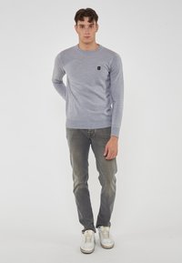 Maglione grigio lavorato a maglia con scollo rotondo, maniche lunghe e una piccola toppa con logo sul petto; abbinato a jeans grigi chiari e sneakers bianche.
