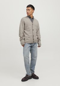 Cardigan lavorato a maglia grigio chiaro, abbottonato, sopra una camicia a quadri bianca e nera. Indossato con jeans blu e scarpe scure. Taglio semplice e rilassato.
