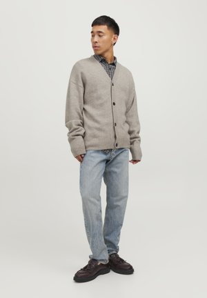Cardigan lavorato a maglia grigio chiaro, abbottonato, sopra una camicia a quadri bianca e nera. Indossato con jeans blu e scarpe scure. Taglio semplice e rilassato.