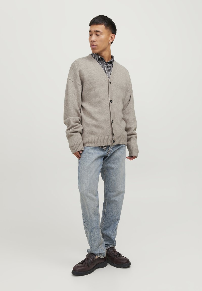 Cardigan lavorato a maglia grigio chiaro, abbottonato, sopra una camicia a quadri bianca e nera. Indossato con jeans blu e scarpe scure. Taglio semplice e rilassato.