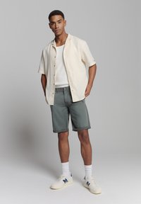 Korte mouwen crèmekleurig overhemd met knopen, witte tanktop, groene shorts en witte sneakers met marineblauwe accenten, tegen een neutrale achtergrond.