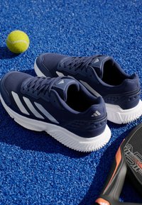 Granatowe buty sportowe z teksturowaną cholewką, białymi akcentami, rowkowanymi podeszwami oraz wyraźnym logo, umieszczone na niebieskiej teksturowanej powierzchni.