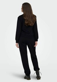 Γυναίκα με μακριά καστανά μαλλιά, φορώντας μαύρη φούτερ και παντελόνι, που στέκεται με την πλάτη στραμμένη προς τα πίσω, με μαύρες δερμάτινες μποτόνες, μπροστά από ένα απλό φόντο.