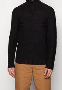 Pull à col roulé noir avec texture côtelée, coupe ajustée et manches longues. Présenté avec un pantalon beige mettant en valeur des détails de quincaillerie visibles.