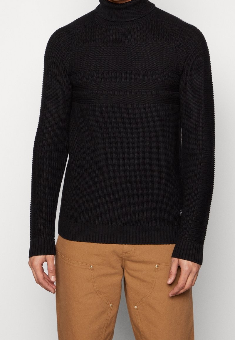 Pull à col roulé noir avec texture côtelée, coupe ajustée et manches longues. Présenté avec un pantalon beige mettant en valeur des détails de quincaillerie visibles.